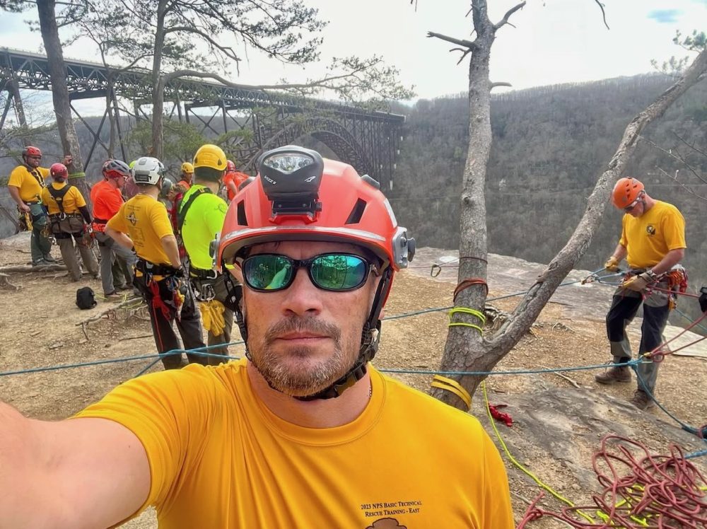 Man wearing a helmet and sunglasses standing near a group of men in the wilderness
