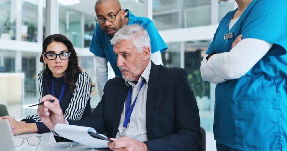 Two health professionals, dressed in scrubs, look over the shoulders of two health administrators, dressed in business attire, to observe the screen of one of the administrator's laptop.