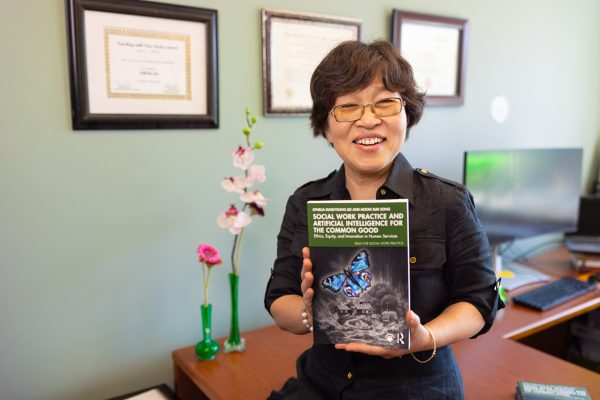 A person with short hair and glasses smiles while holding a book titled "Social Work Practice and Artificial Intelligence for the Common Good" in an office setting.