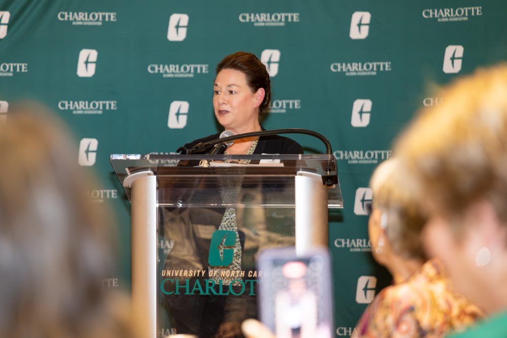 A person speaks at a podium during an event hosted by the Charlotte Alumni Association, with attendees in the foreground.