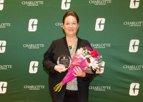 A person stands in front of a green backdrop with the Charlotte Alumni Association logo, holding a glass award and a bouquet of flowers.